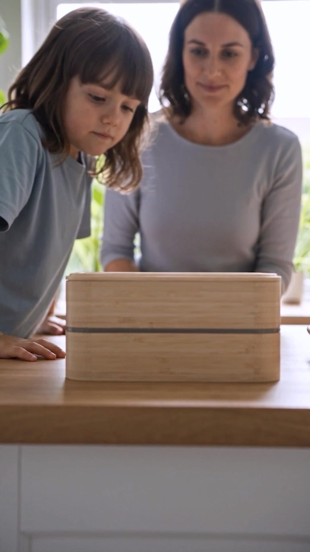 Una fiambrera de madera en primer término sobre la encimera, con una madre y su hija detrás.